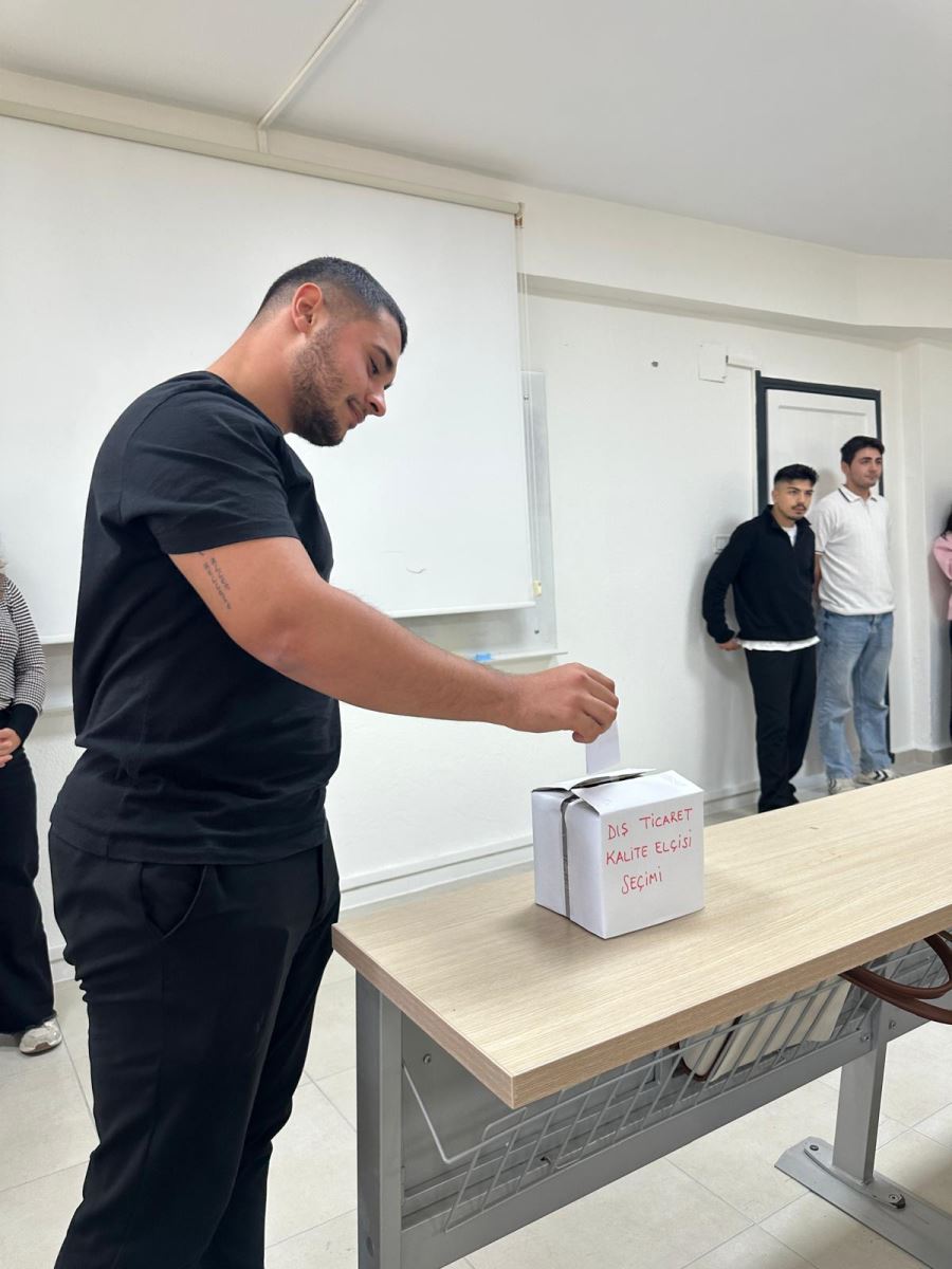 A male student is seen voting in a classroom election.  * He is wearing a black t-shirt and black pants, placing a piece of paper into a white box labeled “Foreign Trade Quality Ambassador Election.”  * The box sits on a light-colored wooden desk.  * In the background, several students stand near the wall watching the process. The scene captures the atmosphere of a small classroom election event.